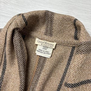 May include: Close-up of a tan and brown wool jacket with black stripes. The jacket has a label that reads "Gianni Versace Made in Italy". The fabric is textured and appears to be a high-quality material.