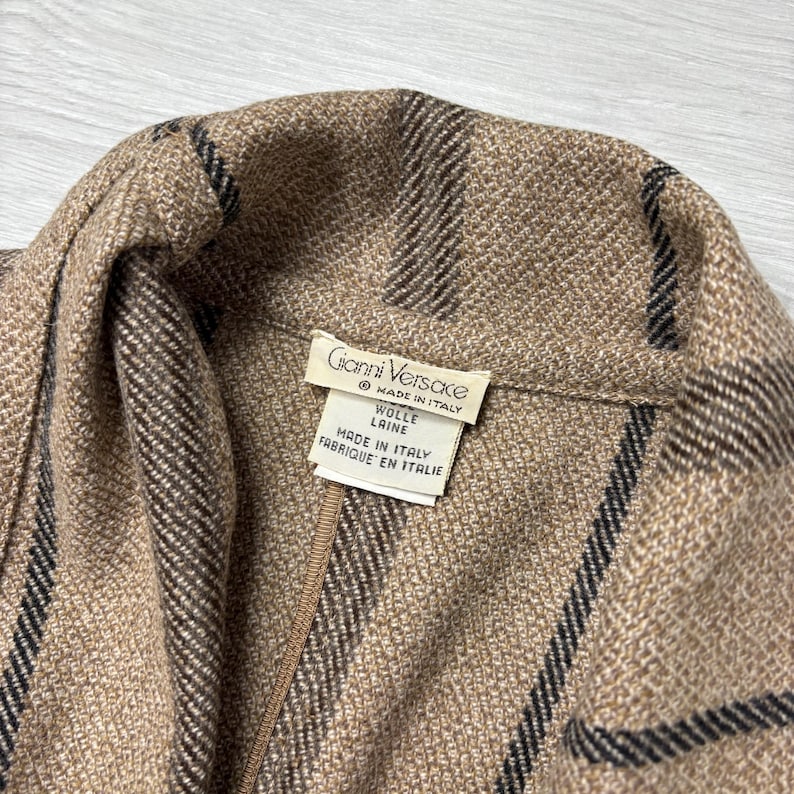 May include: Close-up of a tan and brown wool jacket with black stripes. The jacket has a label that reads "Gianni Versace Made in Italy". The fabric is textured and appears to be a high-quality material.