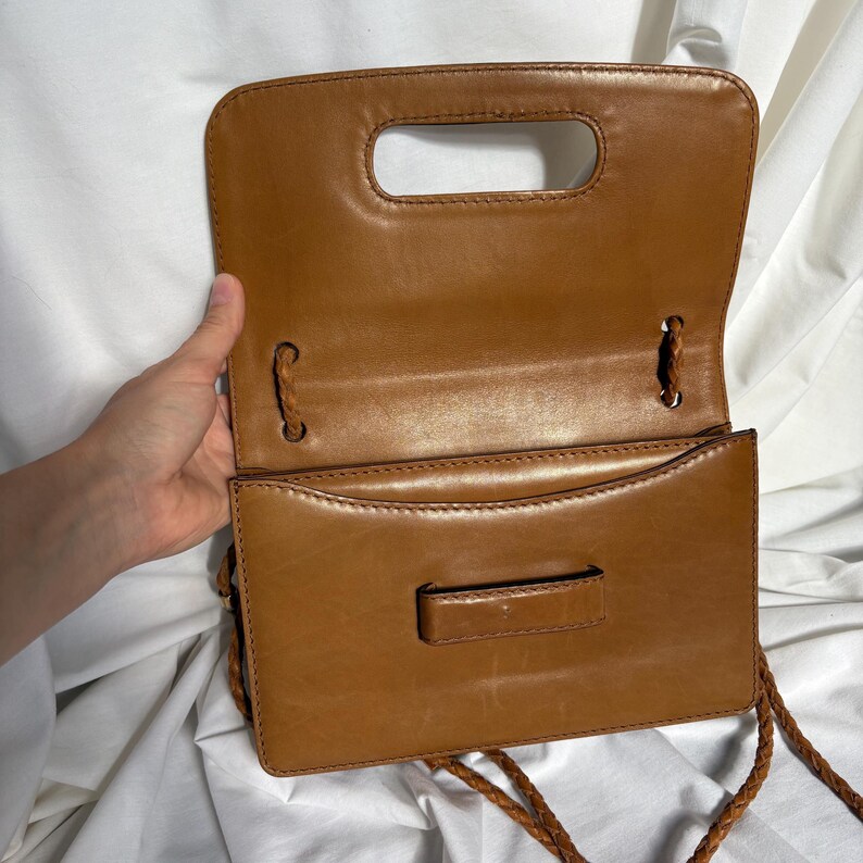 May include: Open, tan-colored leather handbag with a handle and a braided shoulder strap. The bag has a rectangular shape with a flap closure and a small, rectangular clasp. The interior is visible, showing a spacious compartment.