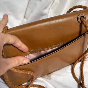 May include: A brown leather crossbody bag with a braided strap. The bag is open, revealing a pink interior. The bag has a rectangular shape and a flap closure. The strap is made of the same brown leather as the bag.