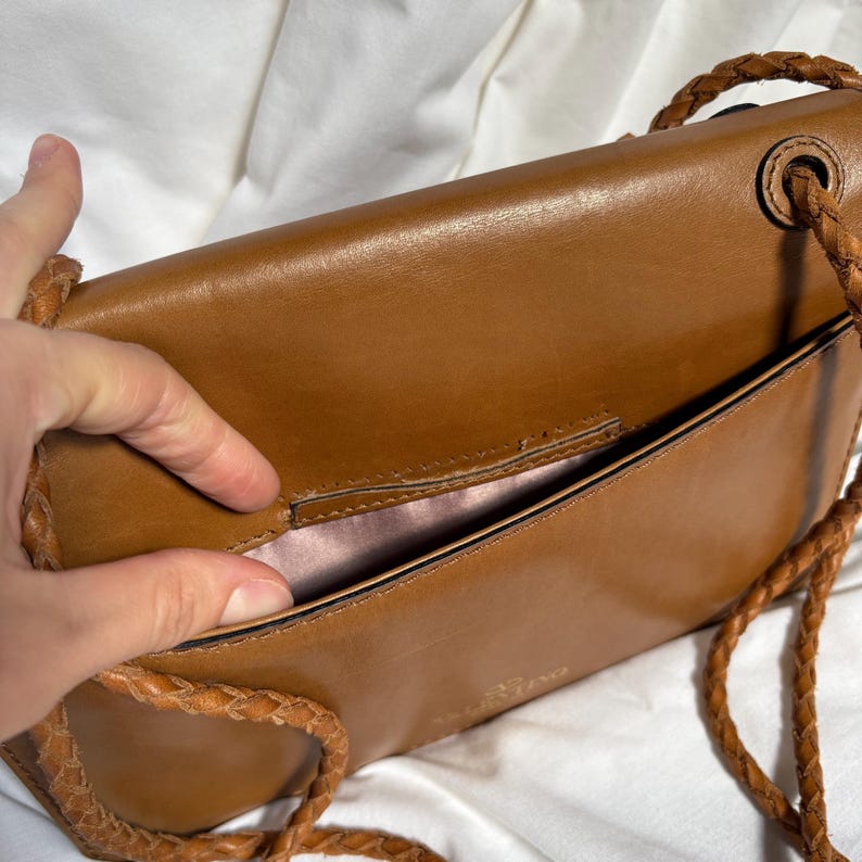 May include: A brown leather crossbody bag with a braided strap. The bag is open, revealing a pink interior. The bag has a rectangular shape and a flap closure. The strap is made of the same brown leather as the bag.