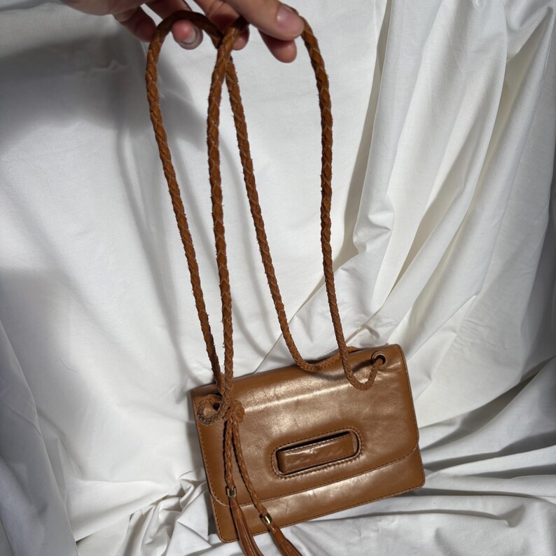 May include: A small, rectangular brown leather handbag with a braided strap. The bag has a rectangular clasp and a tassel detail. The strap is held by a hand.