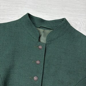 May include: Close-up of a dark green jacket with a mandarin collar and three decorative buttons. The jacket appears to be made of a textured fabric, possibly linen or a similar material. The buttons are a dark, ornate design.