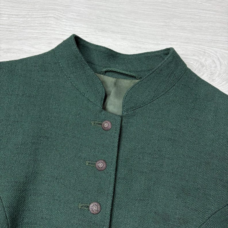 May include: Close-up of a dark green jacket with a mandarin collar and three decorative buttons. The jacket appears to be made of a textured fabric, possibly linen or a similar material. The buttons are a dark, ornate design.