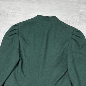 May include: A dark green blazer with a high collar and structured shoulders. The back of the blazer features vertical seams and a tailored fit. The fabric has a subtle texture, and the overall design is classic and elegant.