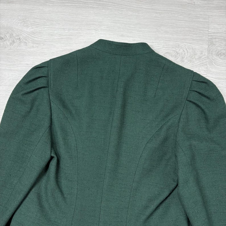 May include: A dark green blazer with a high collar and structured shoulders. The back of the blazer features vertical seams and a tailored fit. The fabric has a subtle texture, and the overall design is classic and elegant.