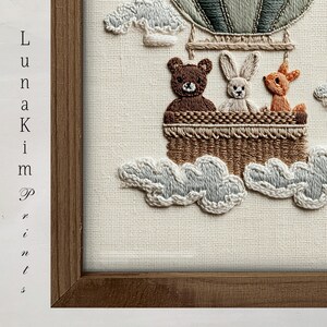 May include: Embroidered artwork featuring a hot air balloon with a bear, bunny, and fox in a woven basket. The balloon is green and tan, with white clouds below. The artwork is framed in a brown wooden frame. The text "Luna Kim Prints" is on the left side.