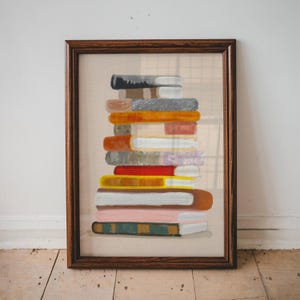Puede incluir: Una obra de arte enmarcada que presenta una pila de libros coloridos. Los libros tienen varios colores, incluyendo naranja, rojo, rosa y verde, con lomos visibles. El marco es de color marrón oscuro y el fondo es beige neutro.