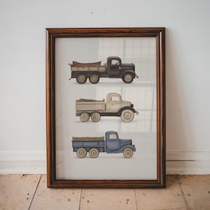 以下が含まれることがあります： 黒、クリーム、青色の3台のヴィンテージピックアップトラックが描かれたフレームプリント。トラックは詳細にイラストされ、ニュートラルな背景に配置され、濃い色の木材でフレームされています。フレームは約30cm x 40cmです。