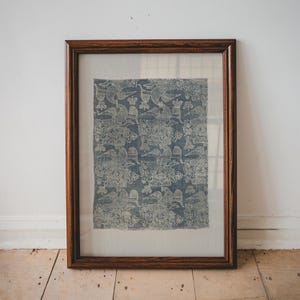 May include: A framed textile art piece featuring a blue and white patterned design. The artwork is enclosed in a dark brown wooden frame, adding a classic touch. The textile has a repeating pattern of birds and floral elements.