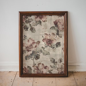 May include: A framed floral print featuring large, muted pink and purple flowers with green leaves against a light beige background. The print is enclosed in a dark brown wooden frame, creating a vintage aesthetic. The artwork is leaning against a white wall.