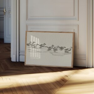 May include: A framed print of a black and white illustration of a flock of ducks swimming in a line. The print is on a light beige background and is framed in a natural wood frame.