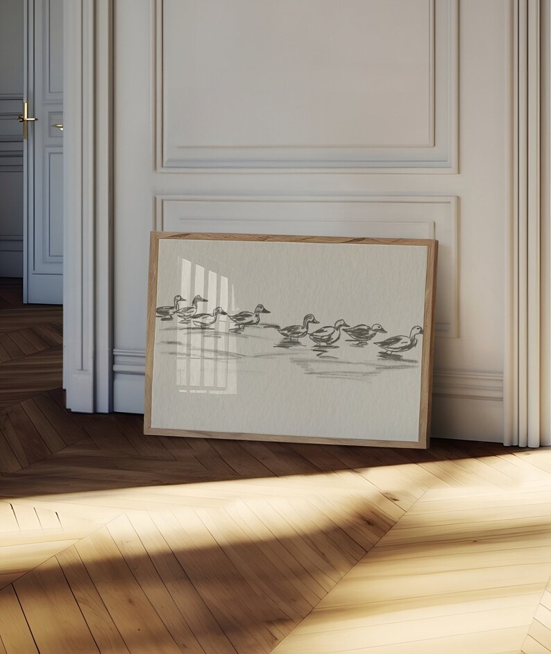 May include: A framed print of a black and white illustration of a flock of ducks swimming in a line. The print is on a light beige background and is framed in a natural wood frame.