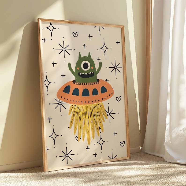 以下が含まれることがあります： オレンジ色のUFOに乗った、一つ目の緑色の宇宙人を描いた額入りのアートプリント。アートワークは、星とハートのイラストが描かれたクリーム色の背景です。宇宙人は歯を見せて笑っており、黄色の光線に囲まれています。