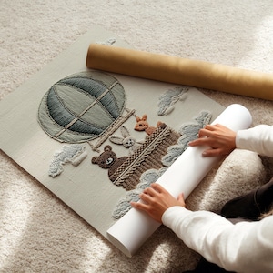 May include: Embroidered artwork featuring a hot air balloon with a bear, rabbit, and deer in a basket. The artwork is in shades of green, brown, and beige. A rolled-up canvas and a cardboard tube are next to the artwork.