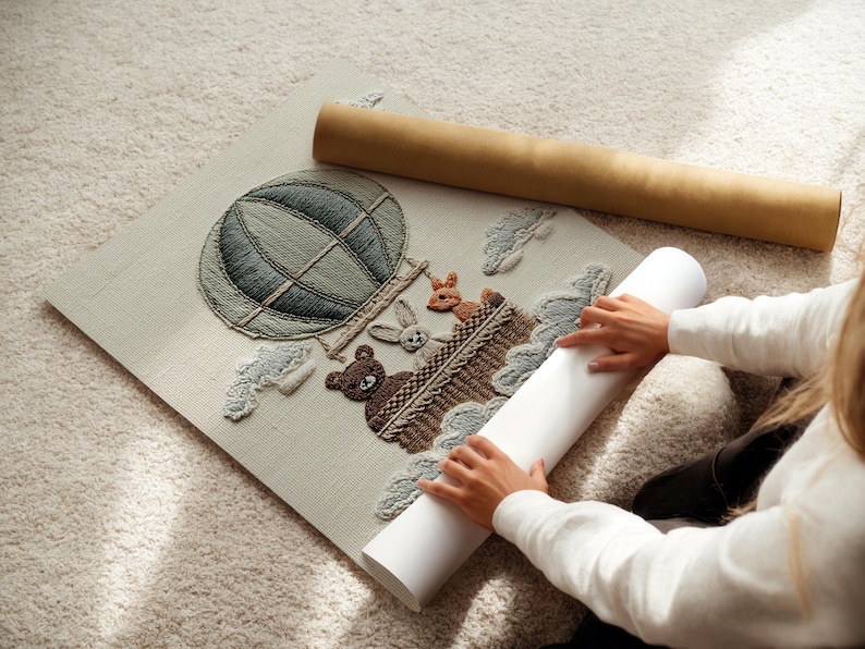 May include: Embroidered artwork featuring a hot air balloon with a bear, rabbit, and deer in a basket. The artwork is in shades of green, brown, and beige. A rolled-up canvas and a cardboard tube are next to the artwork.