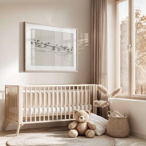 May include: A light wood crib with a white mattress and a beige and white striped fitted sheet. A brown teddy bear sits on the floor in front of the crib. A wicker basket with a white blanket sits on the floor next to the crib. A large framed print of a flock of birds in flight hangs on the wall above the crib.