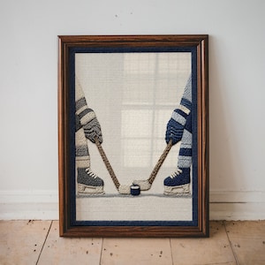 May include: A framed needlepoint art piece depicting two hockey players facing off. The players are shown from the waist down, wearing skates and socks in shades of blue, white, and grey, each holding a wooden hockey stick toward a black puck.