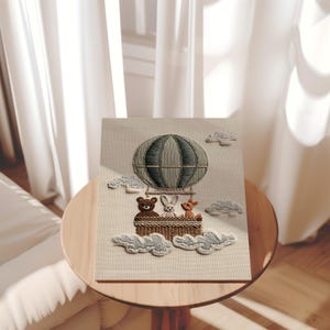 May include: Embroidered artwork featuring a hot air balloon with a gray and blue striped design. The basket holds a brown bear, a white rabbit, and a brown deer. The artwork includes embroidered clouds and is displayed on a wooden table.