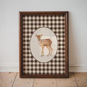 May include: A framed artwork featuring a watercolour painting of a fawn with white spots, set against a brown and white checked background. The frame is a dark wood colour. The artwork is displayed on a white wall and wooden floor.
