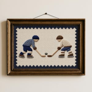 May include: A framed embroidery piece featuring two hockey players in blue and beige uniforms facing each other on a cream-colored fabric background. The players are positioned with hockey sticks near a black puck, surrounded by a dark blue zigzag border.
