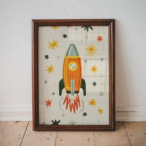 Op de afbeelding: Een ingelijste print met een kleurrijke illustratie van een raket. De oranje raket heeft een blauwe bovenkant en zwarte vinnen, met een rond venster. De achtergrond is licht met gele, rode en zwarte sterren.