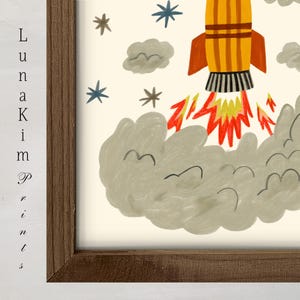 May include: A framed art print featuring a yellow rocket ship with orange fins launching into space, surrounded by gray clouds, red and yellow flames, and blue and brown stars. The text "Luna Kim Prints" is visible on the left side of the frame.