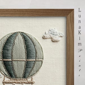 May include: Embroidered artwork featuring a hot air balloon in shades of green and beige, with a fluffy cloud. The artwork is framed in a natural wood frame and set against a neutral fabric background. The text "Luna Kim Prints" is visible.