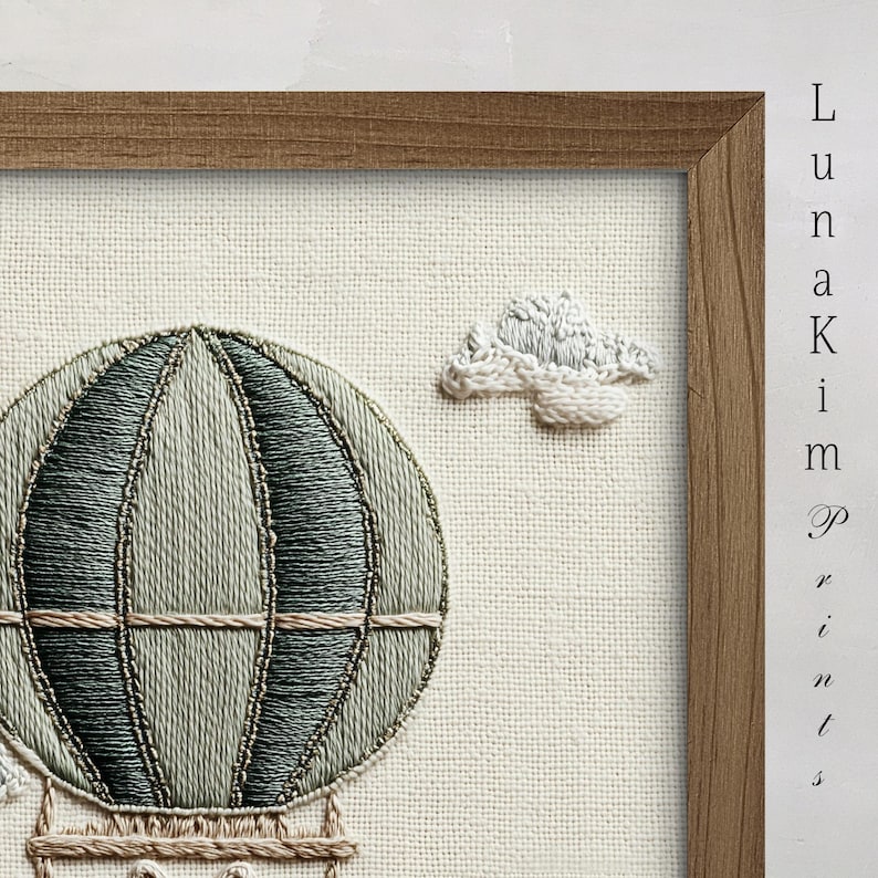 May include: Embroidered artwork featuring a hot air balloon in shades of green and beige, with a fluffy cloud. The artwork is framed in a natural wood frame and set against a neutral fabric background. The text "Luna Kim Prints" is visible.