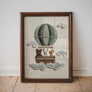 May include: Embroidered artwork in a wooden frame. The image depicts a hot air balloon with a basket holding a bear, rabbit, and deer. The balloon is gray and the clouds are light gray. The artwork is on a cream-colored background.