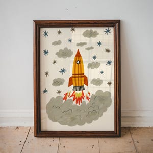 May include: A framed wall art print featuring a yellow and orange rocket ship launching into a sky filled with gray clouds and blue and brown stars. The rocket has a pointed nose, a circular window, and red and orange flames at the base.