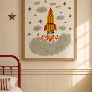 May include: A framed wall art print featuring a yellow and orange rocket ship launching into space. The illustration includes grey clouds, blue and brown stars, and a large grey smoke plume at the base. The artwork is enclosed in a light wood frame.