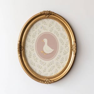 Puede incluir: Un marco ovalado dorado con una ilustraci&oacute;n de pato blanco sobre fondo rosa. El fondo de la imagen es una pared blanca con un patr&oacute;n floral claro.