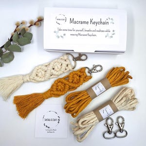 Puede incluir: Un kit de llavero macramé con dos llaveros terminados, uno crema y otro amarillo mostaza, y dos madejas de cordón a juego. El kit incluye dos mosquetones plateados y una caja con el texto "Macrame Keychain".