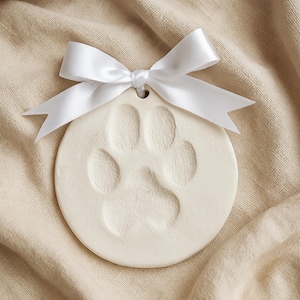 May include: A circular, off-white ceramic ornament with a paw print. A white satin ribbon is tied in a bow at the top. The ornament is displayed on a beige fabric background.