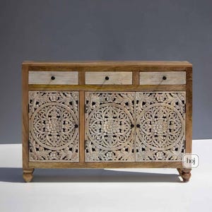 Puede incluir: Un aparador de madera con tres cajones y tres puertas. Los cajones son de color claro con pomos negros. Las puertas presentan intrincados diseños florales tallados. El aparador tiene un acabado de madera natural y está sostenido por patas cortas y redondeadas.