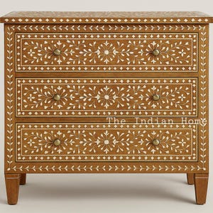 Op de afbeelding: Een houten dressoir met drie lades en ingewikkelde witte bloemeninleg. De kast heeft messing knoppen en de tekst "The Indian Home" op de voorkant. Het hout heeft een warme, bruine tint en staat op korte poten.