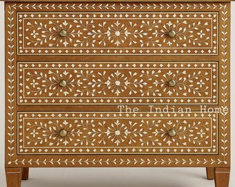 May include: A three-drawer wooden chest with intricate white floral inlay designs. The chest features brass knobs and the text "The Indian Home" on the front. The wood has a warm, brown tone, and the chest sits on short legs.