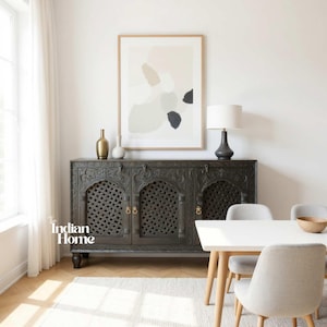 May include: A dark grey carved wooden cabinet with four doors and brass handles. Above the cabinet is a framed abstract art print with neutral colours. A white table and chairs are in front of the cabinet. A lamp and vases are on top.