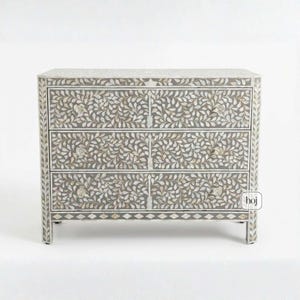 May include: A grey chest of drawers with four drawers, featuring a white leaf design. The chest has a rectangular shape with a decorative border and crystal knobs. The design is elegant, suitable for bedroom or living room decor. The chest is approximately 100cm wide.