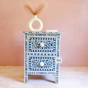 Könnte beinhalten: Ein blau-weißes Nachttischchen mit floralem Muster und zwei Schubladen. Auf dem Nachttisch steht eine weiße Vase mit dekorativem Gras. Der Tisch ist aus Holz gefertigt und hat ein Blumendesign auf der Vorderseite.