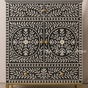 Könnte beinhalten: Ein schwarz-weißes Schränkchen mit aufwendigen floralen Mustern und goldenen Akzenten. Das Schränkchen hat zwei Türen und zwei Schubladen mit goldenen Knöpfen. Das Schränkchen ist aus Holz und hat einen traditionellen Stil.