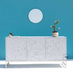Könnte beinhalten: Weißes Sideboard mit floralem Muster auf Türen und Schubladen, vor einer türkisfarbenen Wand. Der Schrank hat ein silbernes Metallgestell und ist mit einer kleinen Pflanze, einer Tasse und einem runden Spiegel versehen. Ca. 150 cm lang.