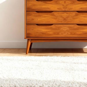 A handcrafted mid-century modern teak wood dresser with spacious drawers, featuring elegant curved drawer fronts and tapered legs. The dresser is made from solid natural teak with rich warm grains, placed in a bright minimalist room with white walls, soft natural light, a framed abstract artwork, two ceramic vases, and a textured white rug on light wood flooring. Modern, Scandinavian, and Japandi-inspired wooden furniture for premium home interiors.