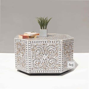 Peut inclure: Table basse octogonale avec un motif floral incrusté. La table présente une base gris foncé et un dessus orné de motifs de feuilles blanches et dorées. Une petite plante en pot et des livres reposent sur la surface de la table.
