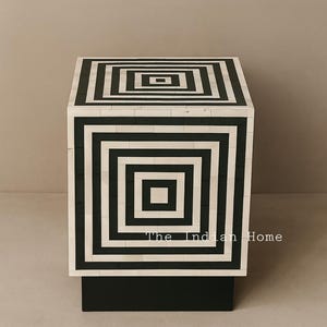 Può includere: Un tavolino con motivo geometrico in bianco e nero. Il tavolo a forma di cubo presenta un motivo quadrato ripetuto sulla parte superiore e sui lati. La base è nera e il testo "The Indian Home" è visibile sulla parte anteriore.