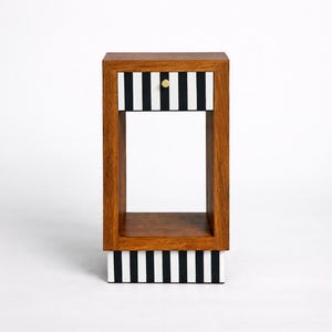 Può includere: Tavolino in legno con cornice rettangolare e cassetto. Il cassetto e la base presentano strisce verticali bianche e nere. Il cassetto ha un piccolo pomello dorato. Il tavolo è alto circa 51 cm.