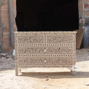 Puede incluir: Cómoda marrón y crema con intrincados diseños de incrustaciones florales y geométricas. La cómoda tiene cuatro cajones y forma rectangular, con patas decorativas. El estilo es ornamentado y detallado, con una altura aproximada de 80 cm.