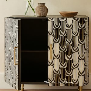 May include: A black and gold cabinet with a geometric pattern, open to reveal a black interior with a shelf. The cabinet has gold-coloured handles and legs. A vase with green leaves and a small brown vase sit on top.