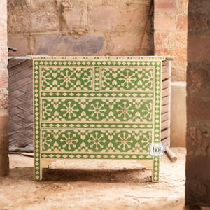 Könnte beinhalten: Eine Holzkommode mit einem geometrischen Muster in Grün und Creme. Die Kommode hat vier Schubladen und ein oberes Fach. Das Design zeigt Blumen- und Rautenformen. Die Kommode steht vor einer Backsteinmauer.